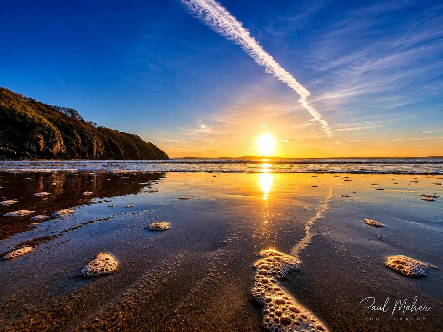 Sun low over a mirror-smooth strand at low tide with a contrail above