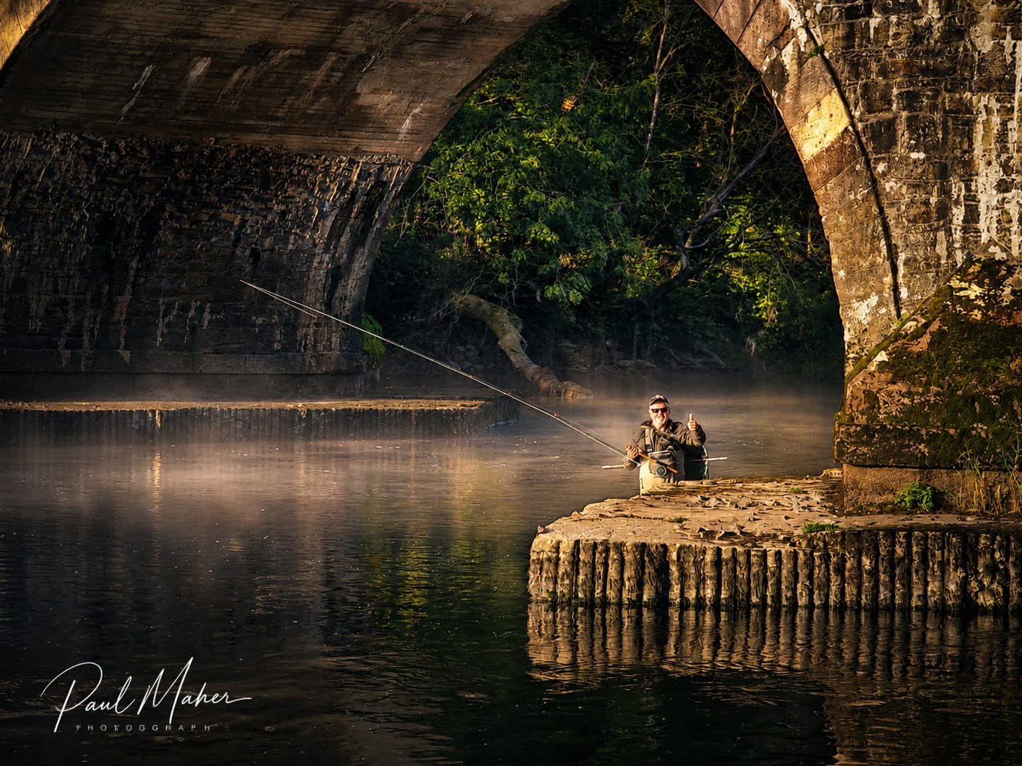An angler fishing beneath a stone bridge arch on a still morning