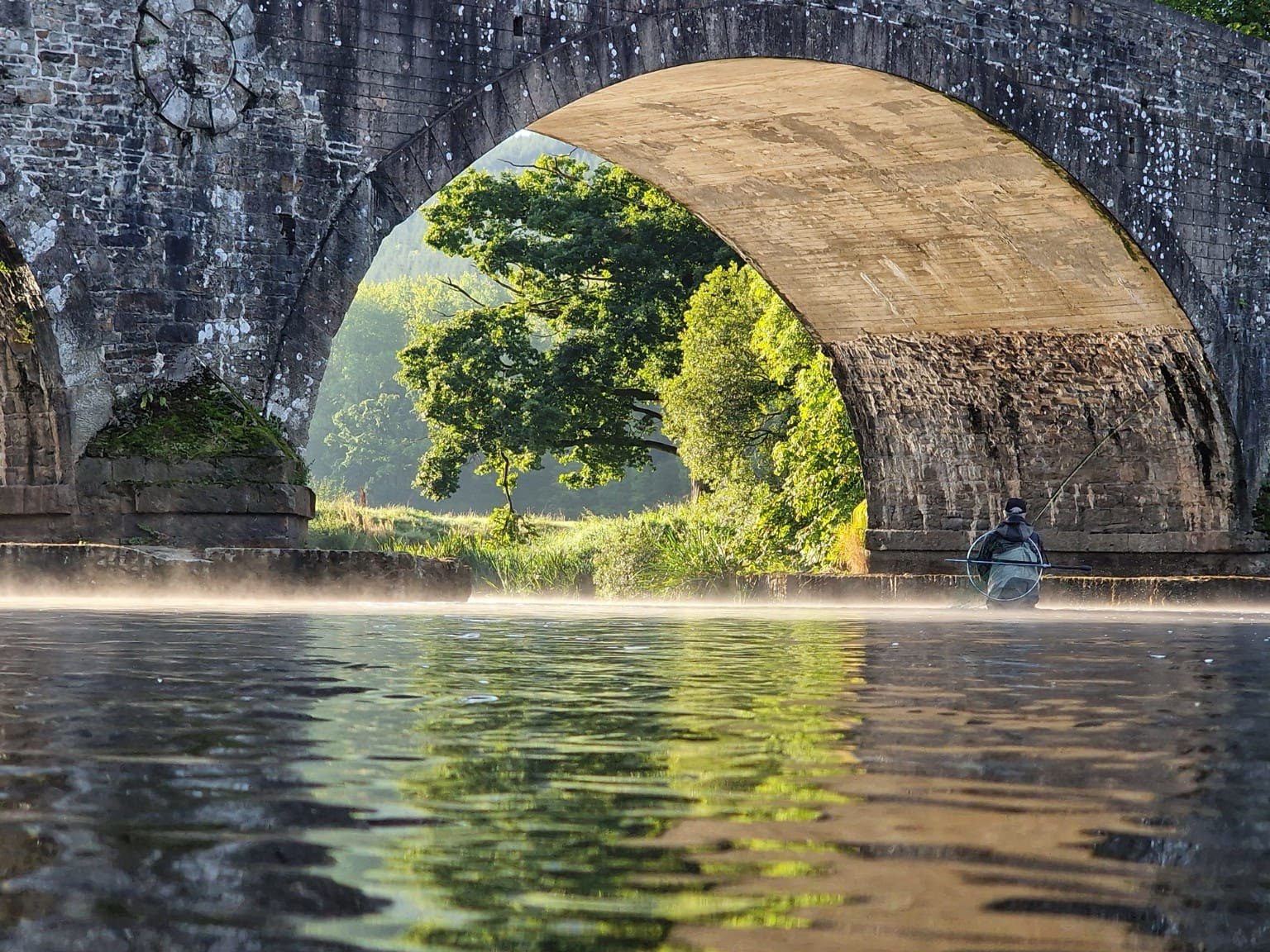 A stone bridge arch spanning quiet water framed by green trees